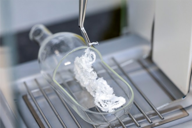 Tweezers holding white material inside a glass container on a metal rack.