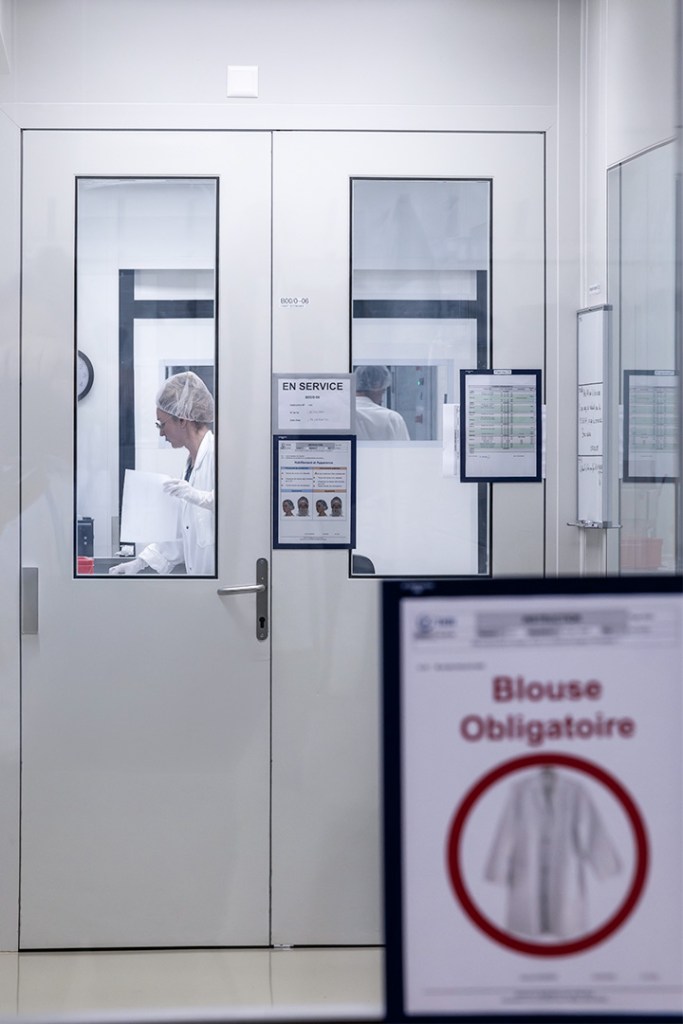 A person in a lab coat and hairnet working inside a laboratory room.