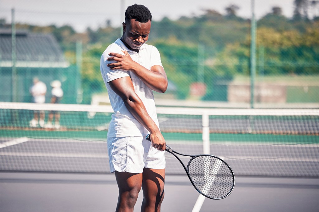 Man in white sportswear holding a tennis racket, grimacing while touching his shoulder.